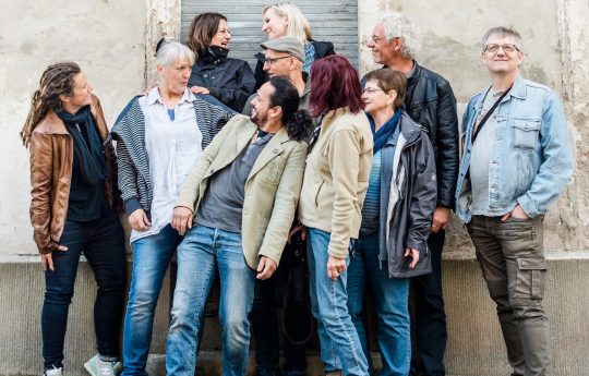 Gruppenfoto diverse Menschen mittleren Alters vor Hauswand lachen und sprechen miteinander